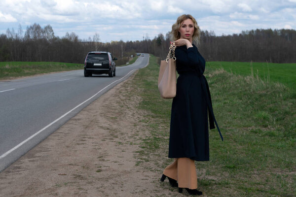 A middle-aged Caucasian woman in an elegant summer black coat stands alone near a highway in the countryside against a cloudy sky on a spring day. Copy space for your text or design.