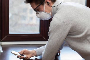 Tıbbi maskeli genç bir adam evdeki karantina sırasında telefonla selfie çekiyor.