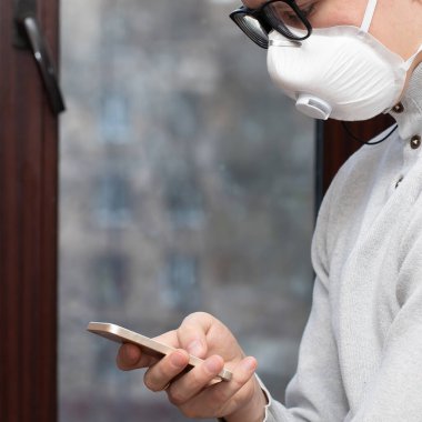 Tıbbi maskeli genç bir adam evdeki karantina sırasında telefonla selfie çekiyor.