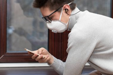 Tıbbi maskeli genç bir adam evdeki karantina sırasında telefonla selfie çekiyor.