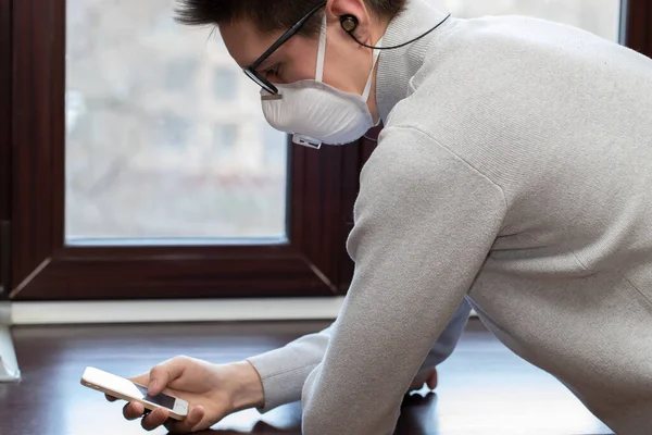 Tıbbi maskeli genç bir adam evdeki karantina sırasında telefonla selfie çekiyor.