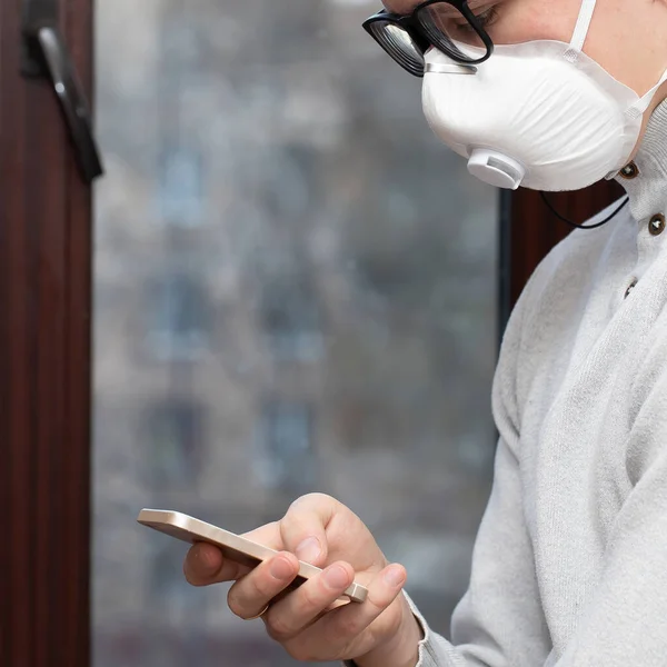 Tıbbi maskeli genç bir adam evdeki karantina sırasında telefonla selfie çekiyor.