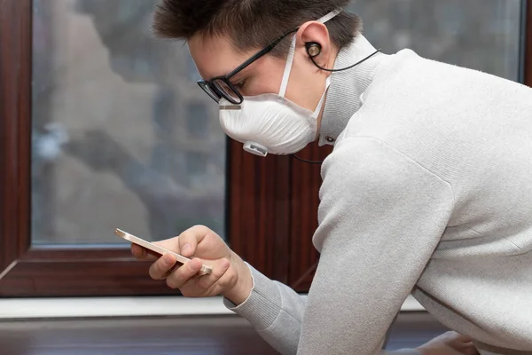 Tıbbi maskeli genç bir adam evdeki karantina sırasında telefonla selfie çekiyor.