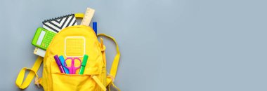 Okula dönmek, eğitim kavramı. Okul malzemeleriyle dolu sarı bir sırt çantası. Not defteri, kalemler, cetvel, hesap makinesi, makas gri arka planda izole edilmiş. Üst Manzara. Boşluğu Kopyala Düz Kompozisyon SancağıName.