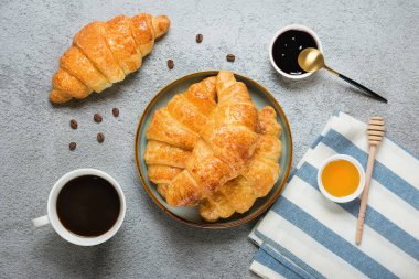 Taze Fransız kruvasanları, tabakta çikolata, yabanmersini reçeli, bal, peçete, beyaz fincan espresso kahvesi, gri granit zemin üzerinde kahve taneleri..