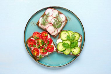 Kahvaltı için çeşitli sandviçler. Ekmek, krem peynir, salatalık, turp, vişne domatesleri, dereotuyla süslenmiş yeşil soğanlar açık mavi masanın üstünde..