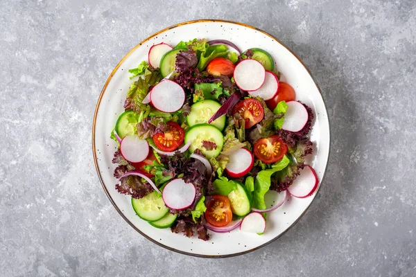 Gezonde groentesalade van kerstomaten, komkommerschijfjes, groene en paarse sla bladeren, uien en olijfolie op bord op betonnen tafel Bovenaanzicht Platte lay Dieet, Mediterraans menu Veganistisch eten — Stockfoto