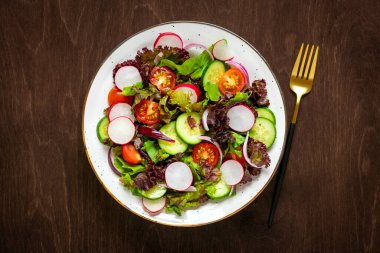 Sağlıklı sebze salatası, kiraz domatesleri, salatalık dilimleri, yeşil ve mor marul yaprakları, soğan ve zeytinyağı tabakta koyu ahşap masa üzerinde.