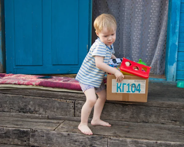 Ранней весной деревня, трехлетний блондин в футболке и трусиках вытаскивает коробку игрушек из дома на улицу. — стоковое фото