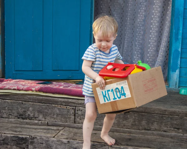 Ранней весной деревня, трехлетний блондин в футболке и трусиках вытаскивает коробку игрушек из дома на улицу. — стоковое фото