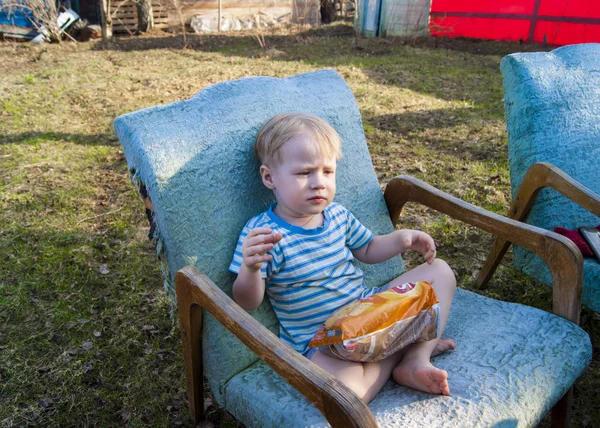 Ранней весной, в деревне, маленький блондин сидит в синем плюшевом кресле на улице во дворе своего дома и ест закуски, на его лице множество эмоций — стоковое фото