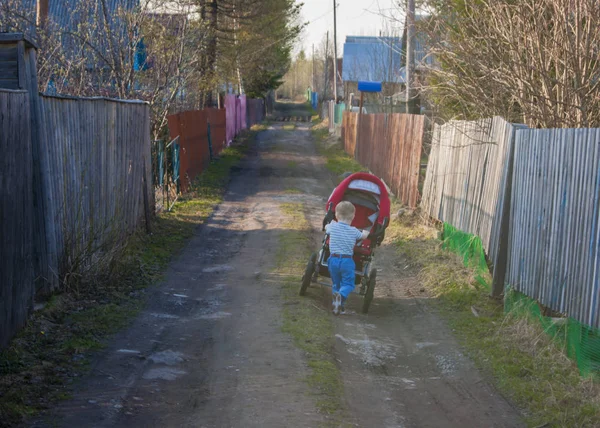 Na jaře, ve vesnici, se malý blonďák sjíždí po vesnické ulici s červeným kočárem — Stock fotografie