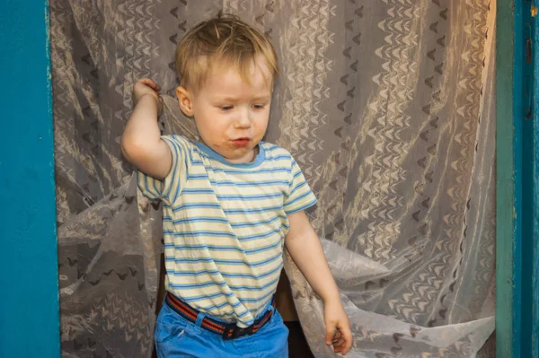 Ранней весной, в деревне, маленький трехлетний блондин Чумазенки выходит из своего деревенского дома, дверь которого покрыта тюлем насекомых. — стоковое фото