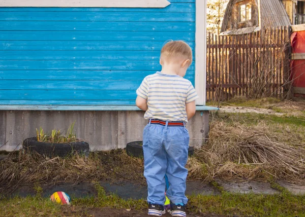 A principios de la primavera, en el pueblo, un niño rubio de tres años de edad, se para con la espalda al público contra la pared azul de su casa de pueblo — Foto de Stock