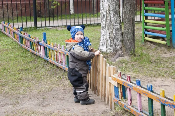 Маленький мальчик трех лет в прохладный весенний солнечный день пытается открыть небольшой деревянный забор, чтобы пойти на игровую площадку детского сада — стоковое фото