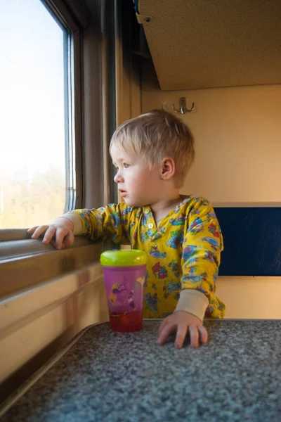 Яркий, милый трехлетний мальчик, едущий на поезде, смотрит в окно, за которым проносится скучный пейзаж. Вертикальное фото . — стоковое фото