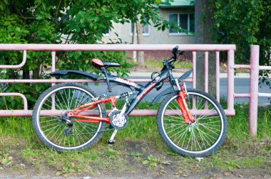 Kırmızı elementli siyah bir bisiklet güneşli bir yaz gününde pembe metal bir çite yaslanır.