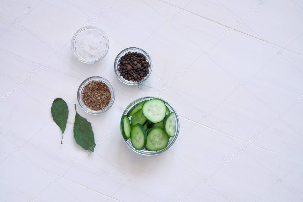 Flat lay composition of fresh cucumber sliced in a glass bowl and the necessary spices for pickling(fermenting)- dill seeds, pepper, salt on a white wooden table.Top view, copy space for text.