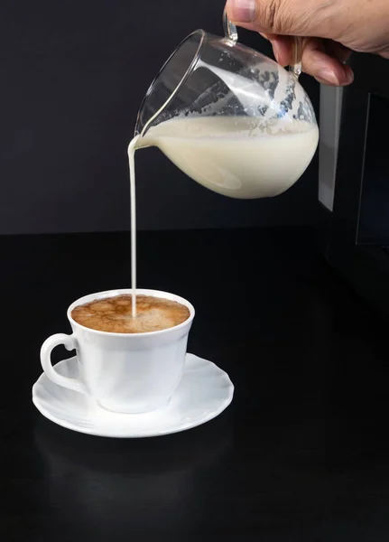 Süt, siyah arka planda köpüklü beyaz bir fincan kahveye dökülüyor. Dikey fotoğraf