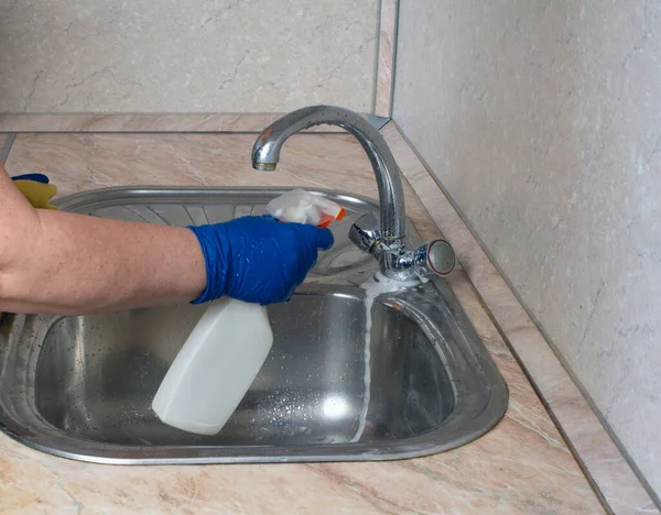 Cleaning the kitchen. A cleaning lady in blue rubber gloves washes a sink with a spray bottle