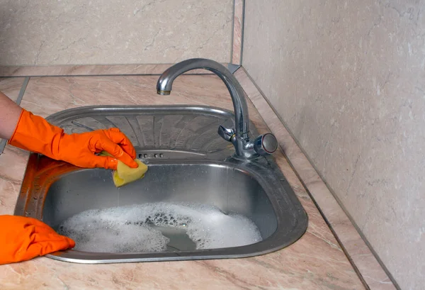 A cleaning lady in orange rubber household gloves washes a sink with a yellow sponge. Soapy water in the sink