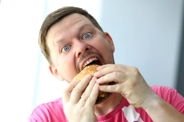 Смешной бородатый мужчина с идиотским выражением лица ест бургер — стоковое фото