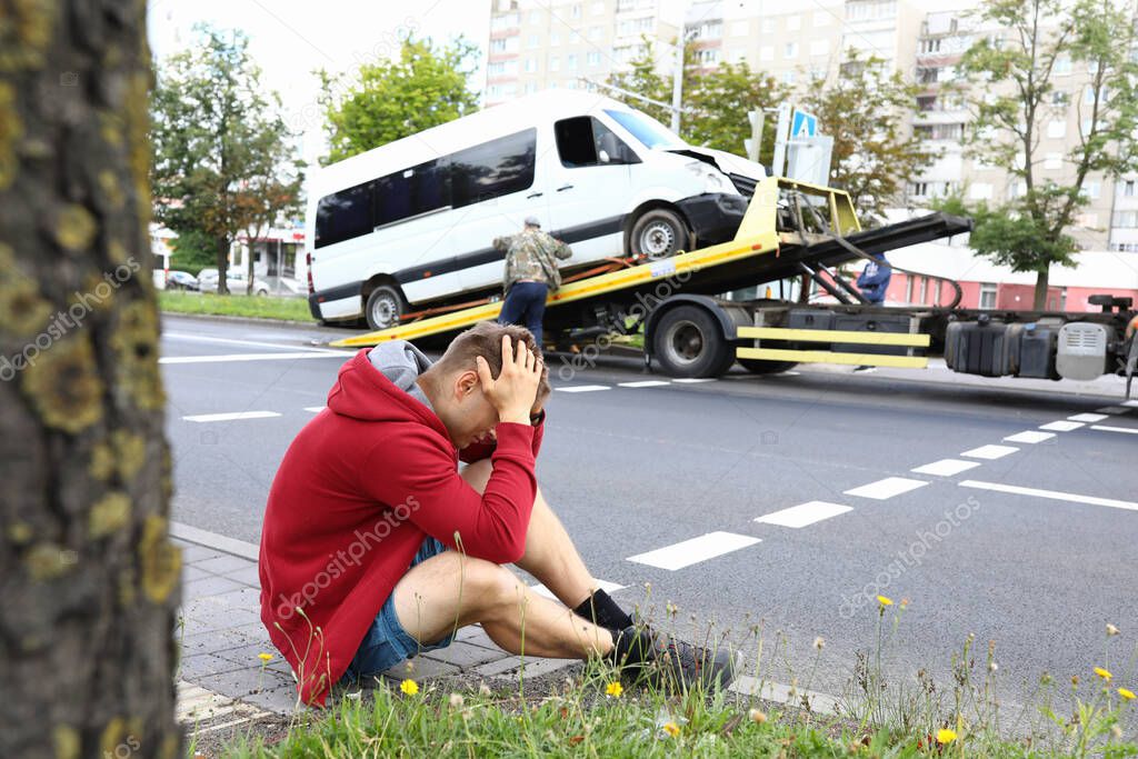 Man sits on side of road holding his head with his hands next to wrecked car after car accident