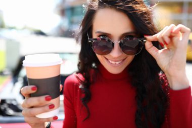 Kadın güneş gözlüğünü çıkaracak ve elinde sıcak bir içecek tutacak.