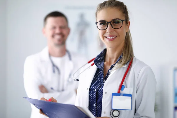 Kvinnlig läkare ler och håller urklipp från bakom hennes kollega står. — Stockfoto