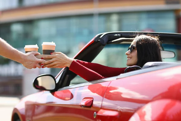 Woman in cabriolet picks up hot drinks from a courier