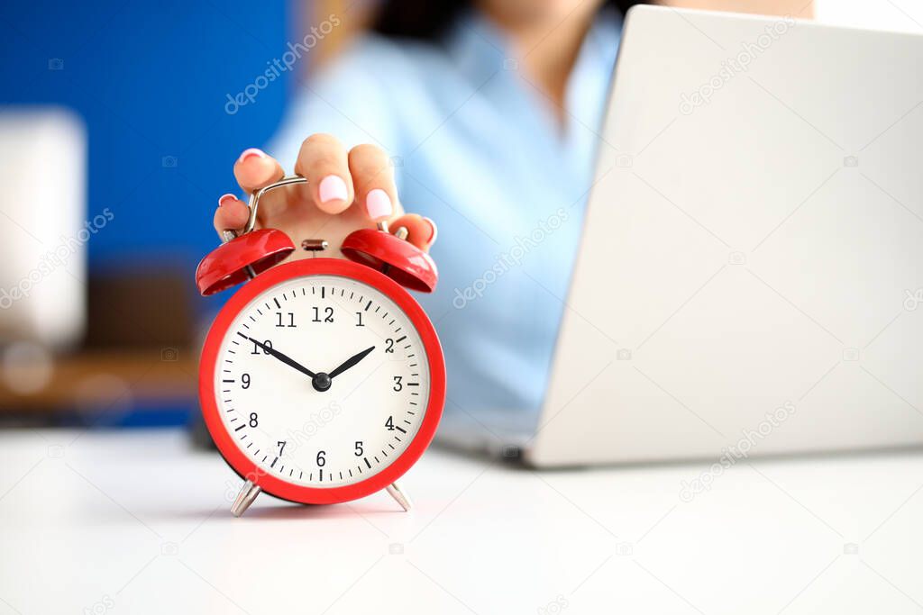 Female hand lies on red alarm clock next to laptop.