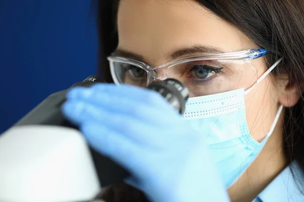 Frau mit Schutzmaske und Handschuhen schaut durchs Mikroskop. — Stockfoto