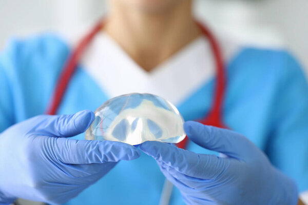 Gloved doctor holds female breast implant in his hands.