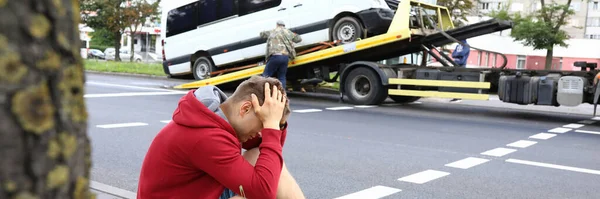 Mężczyzna siedzi na poboczu, trzymając głowę z rękami obok rozbitego samochodu po wypadku samochodowym. — Zdjęcie stockowe