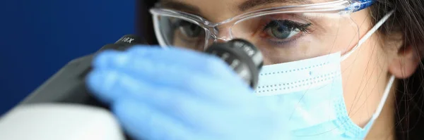 Mulher em máscara protetora e luvas olha através do microscópio . — Fotografia de Stock