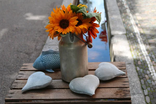 Still life of a bunch of yellow sunflowers with green leaves in a metal vase, three stone hearts and a blue pottery fish on a wooden box