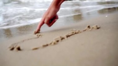 Kadının eli kuma bir kalp çizer ve bir dalga tarafından sürüklenir.