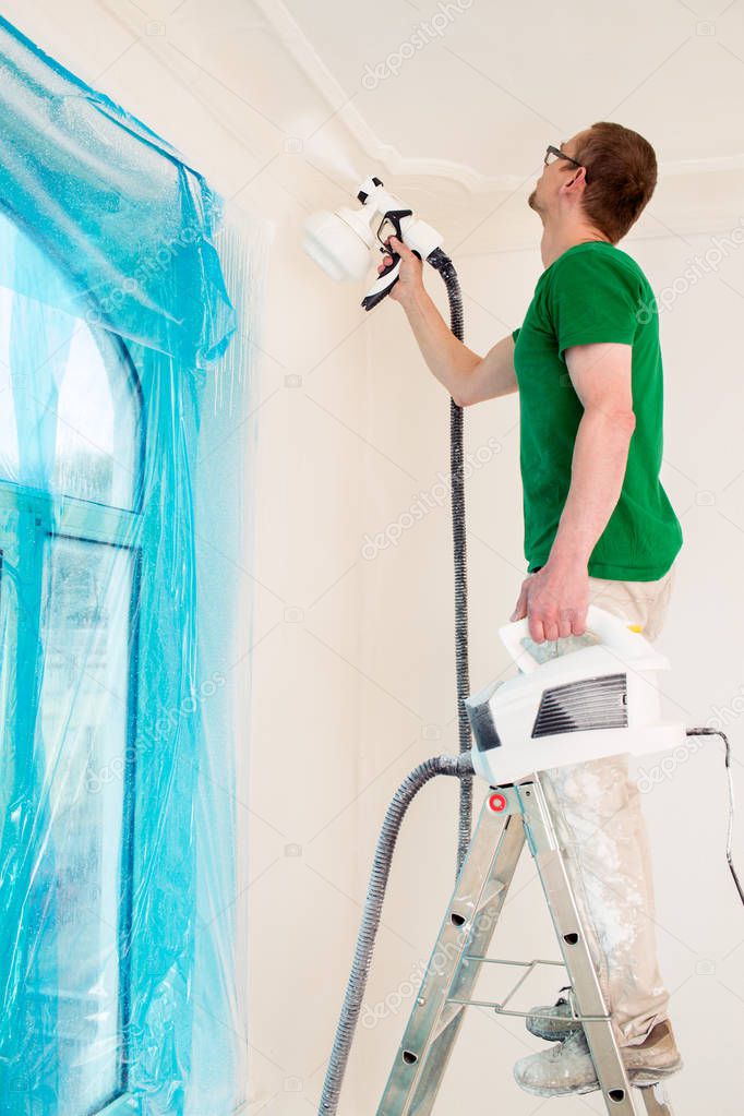 Man painting room walls with paint spray gun while standing on ladder.