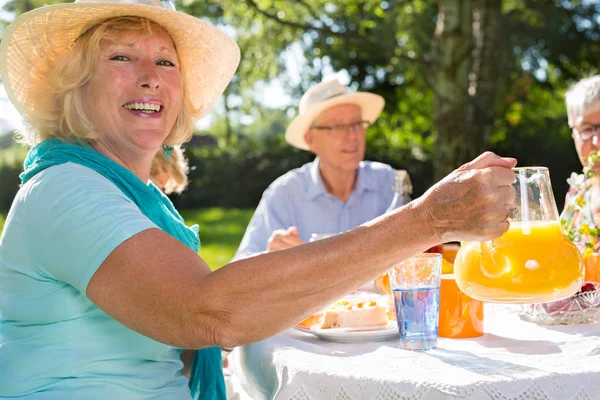 老年人在阳光下户外野餐 金发美女戴帽子 向年迈的朋友提供橙汁 — 图库照片