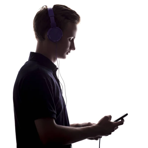 Silhueta de um jovem ouvindo música em fones de ouvido, cara vira através de uma lista de reprodução na tela do smartphone em um fundo isolado branco — Fotografia de Stock