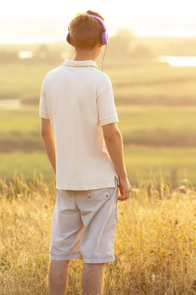 Adolescente em fones de ouvido ouvindo música na natureza, jovem está relaxando e olhando para o pôr do sol no campo de verão — Fotografia de Stock