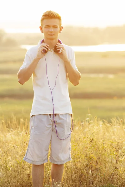 Tinédzser fülhallgató hallgat zenét a természetben, fiatal szőke férfi néz egyenesen a háttérben a gyönyörű nyári táj — Stock Fotó