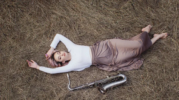 Corps d'une belle jeune femme en robe longue couchée sur l'herbe sèche à côté du saxophone, fille blonde repose sur le foin avec un instrument de musique, vue du dessus — Photo