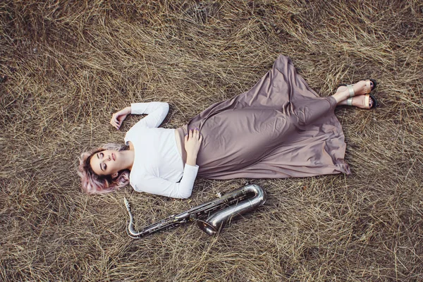 Corps d'une belle jeune femme en robe longue couchée sur l'herbe sèche à côté du saxophone, fille blonde repose sur le foin avec un instrument de musique, vue du dessus — Photo