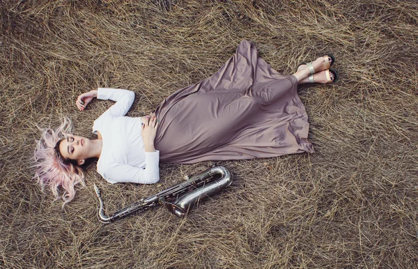 Corps d'une belle jeune femme en robe longue couchée sur l'herbe sèche à côté du saxophone, fille blonde repose sur le foin avec un instrument de musique, vue du dessus — Photo