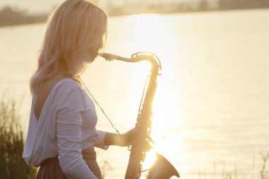 Gün doğumunda nehir kenarında saksafon çalan genç ve güzel bir kızın silueti. Gün doğumunda doğada bir kadın konser veriyor ve rahatlatıcı bir müzik konsepti veriyor.