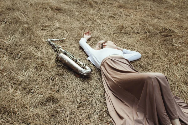 Corps d'une belle jeune femme en robe longue couchée sur l'herbe sèche à côté du saxophone, fille en été sur le foin repose avec un instrument de musique, le concept de détente sur la nature — Photo