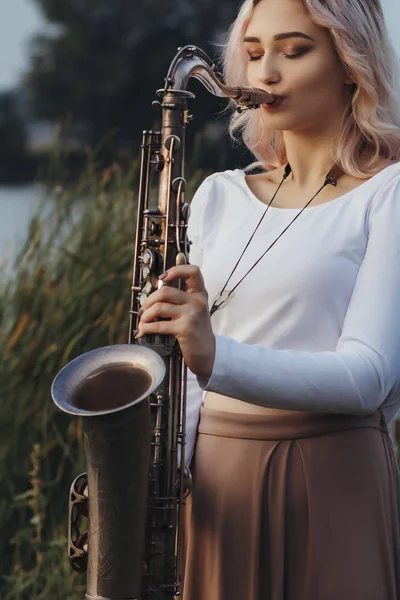 Портрет молодої жінки, яка грає саксофон на березі річки, блондинка з музичним інструментом на світанку, концептуальна музика і розслабтеся — стокове фото