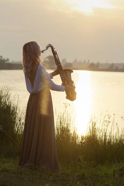 Silueta de una joven hermosa chica tocando el saxofón al amanecer junto al río, una mujer en un vestido largo en la naturaleza al amanecer relajante, música conceptual — Foto de Stock
