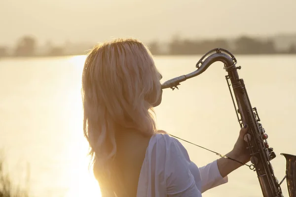 Silhouette d'une jeune belle jeune fille jouant du saxophone au lever du soleil au bord de la rivière, femme sur la nature au lever du soleil donnant un concert et relaxant, concept music — Photo